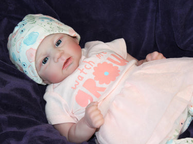 A baby doll dressed in a pink shirt and wearing a hat, sitting against a soft background.